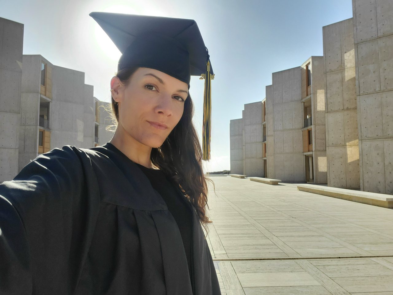 Melanie Kaiser in cap and gown in the Salk Institute courtyard, the concrete towers receding behind her.