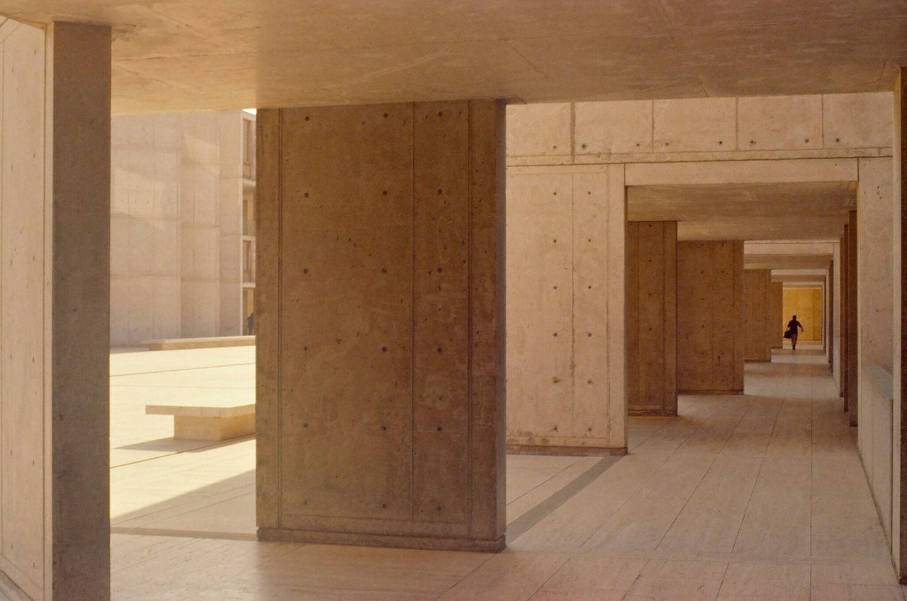 A shaded walkway inside the Salk Institute: concrete piers and travertine paving, a lone figure at the far end.