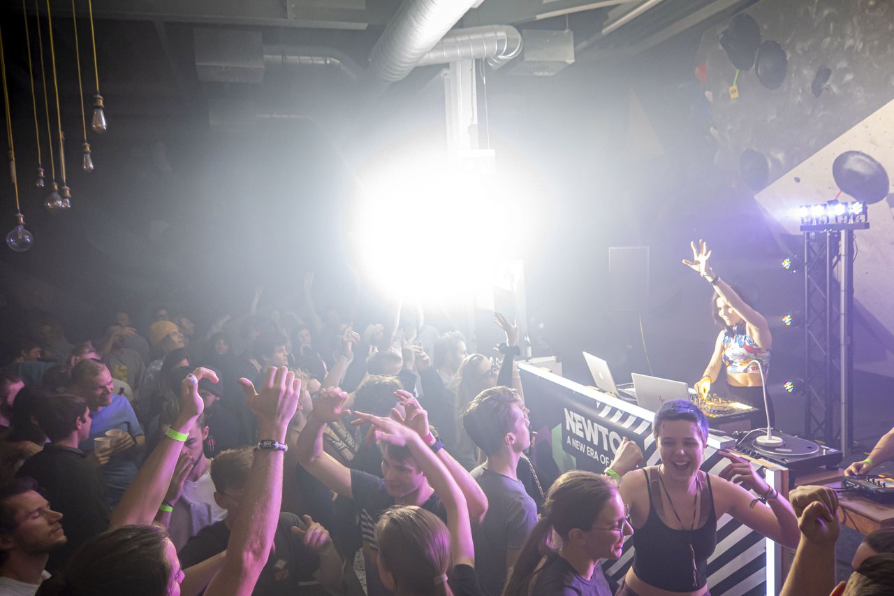 A packed floor of people with their hands up, lit from behind, a DJ raised above the crowd.