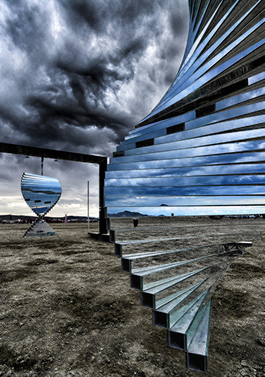 A large sculpture of mirrored, twisting helix forms on a steel frame under a heavy sky, reflecting the landscape around it.