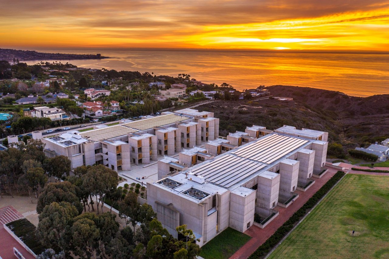 The Salk Institute from the air at sunset: the two mirrored concrete blocks with the courtyard between them, the eucalyptus grove circled along the near edge, and La Jolla and the Pacific beyond with the sun on the horizon.