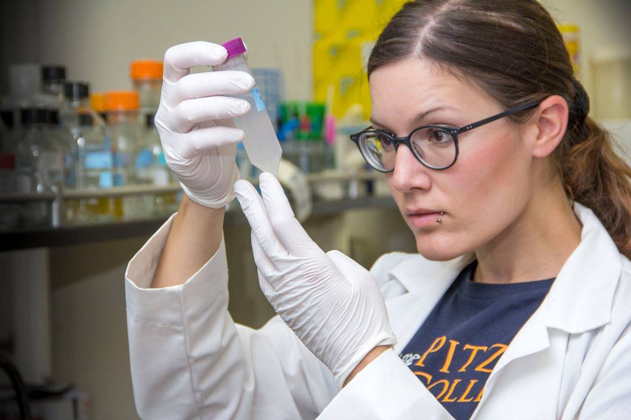 A scientist in a lab coat holding a sample tube up to the light at the bench.